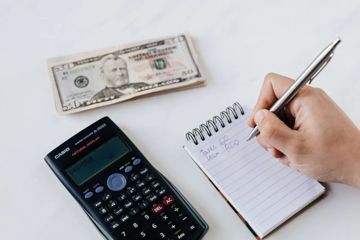 Hand writing expenses in a notepad beside a calculator and banknotes